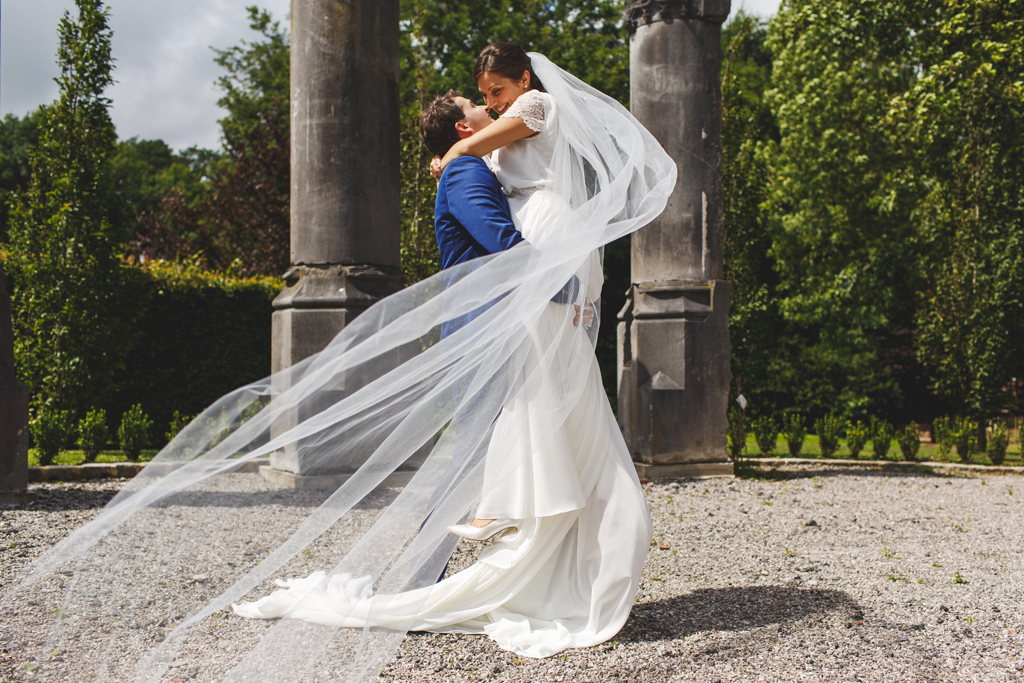 Photo des mariés au château de Morbecque. Séance photo couple sur le domaine. Photographe spécialisé mariage. La mariée est soulevée par le mari et le vent emporte le voile. Robe de mariage Olivier Sinic.